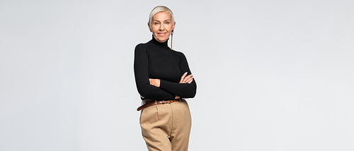 pleased and trendy senior woman in beige pants and turtleneck posing with crossed arms isolated on grey, banner