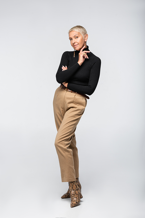full length of pleased and trendy senior woman in beige pants and turtleneck posing on grey
