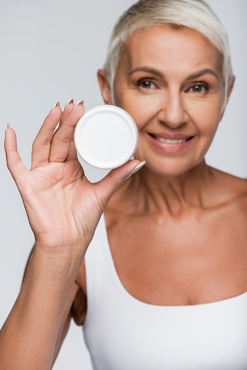 positive elderly woman holding container with cosmetic cream isolated on grey