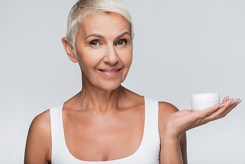happy senior woman showing container with cosmetic cream isolated on grey