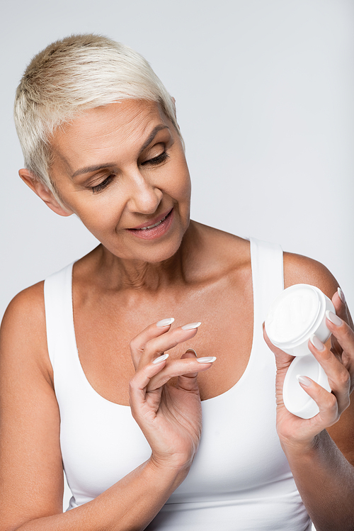 happy senior woman looking at container with face cream isolated on grey