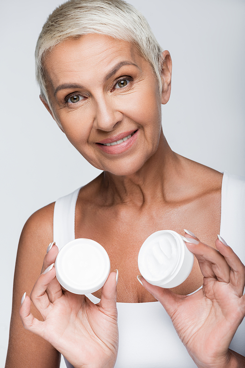 happy senior woman holding container with cosmetic cream isolated on grey