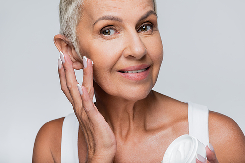 happy senior woman holding container and applying face cream isolated on grey