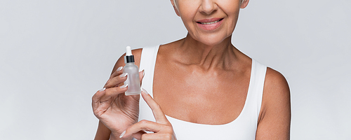 cropped view of happy senior woman holding bottle with serum isolated on grey, banner