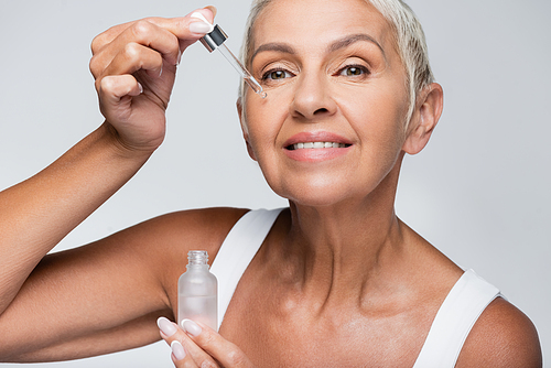 happy senior woman applying vitamin c serum isolated on grey