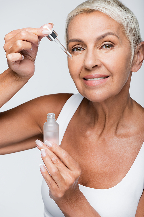 happy senior woman applying serum with pipette isolated on grey