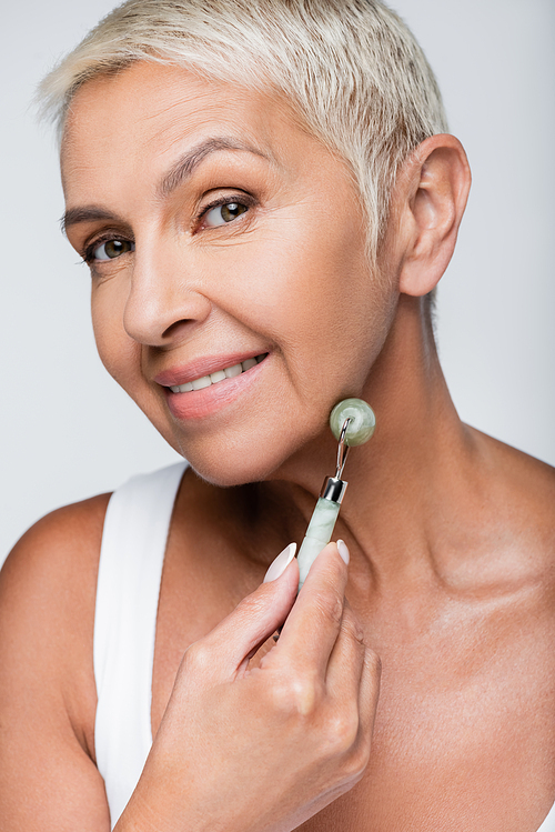 happy senior woman massaging face with jade roller isolated on grey