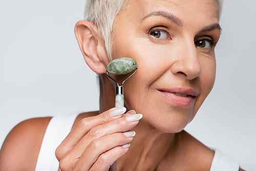 smiling woman with grey hair massaging face with jade roller isolated on grey