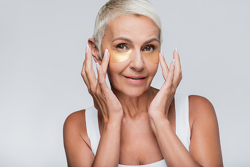 pleased senior woman applying collagen eye patches isolated on grey