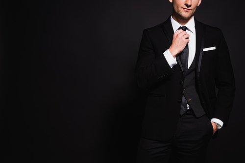partial view of elegant man touching tie while standing with hand in pocket isolated on black