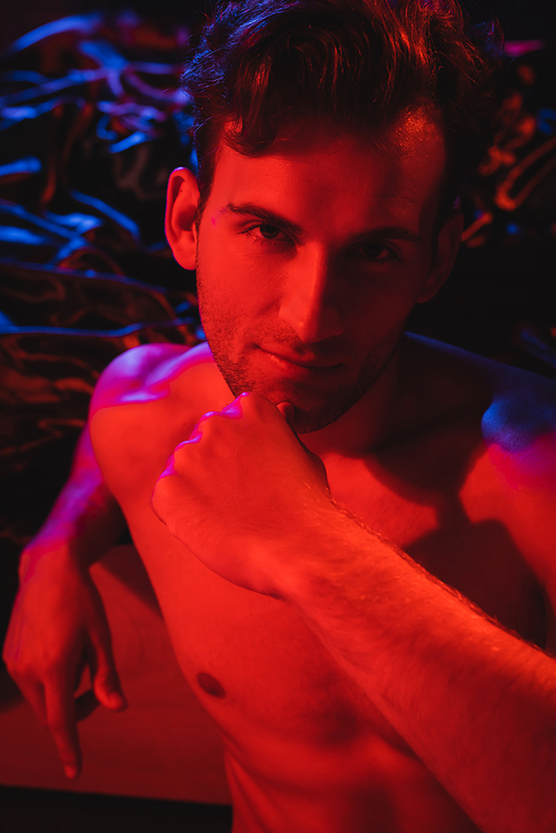 young shirtless man  while posing near black blurred bedding in red lighting