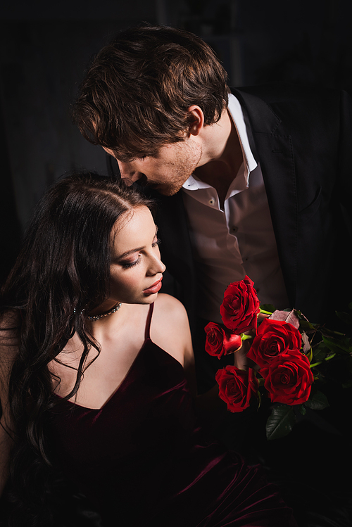 young man holding red roses near elegant brunette woman on dark background