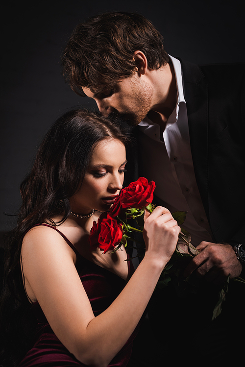 elegant woman smelling red roses near man in suit on dark background