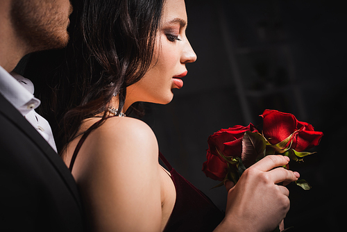 side view of sensual brunette woman holding red roses near young man on dark background
