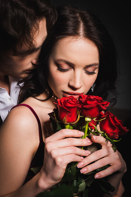 sensual and elegant woman holding red roses near man on dark background