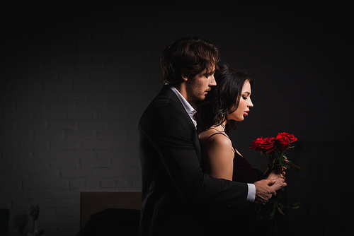 side view of man in suit near sensual brunette woman with red roses