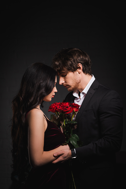 side view of brunette woman with red roses standing face to face with man on dark background