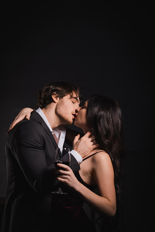 man in black suit kissing elegant woman holding glass of red wine on dark background
