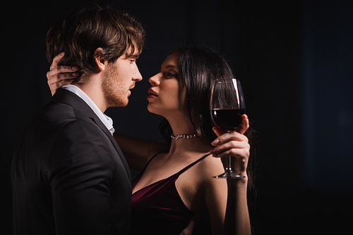elegant woman with glass of red wine and young man in black suit looking at each other on dark background