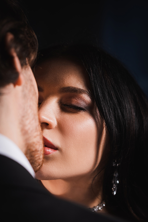 close up view of young brunette woman with closed eyes kissing blurred man on dark background