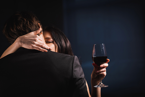back view of man in black suit near woman embracing neck while holding glass of red wine on dark background