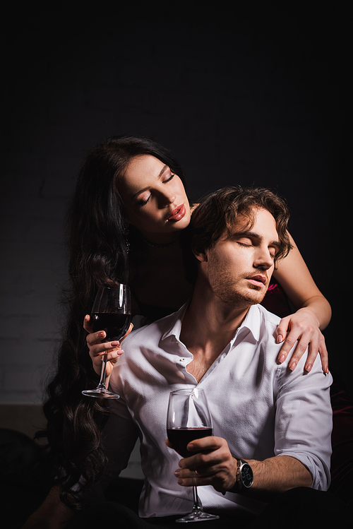 passionate woman with glass of red while seducing young man in white shirt in dark bedroom