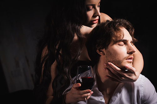 passionate brunette woman holding glass of red wine while embracing neck of man in white shirt on dark background