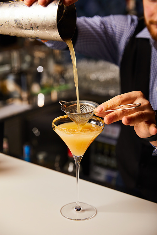 cropped view of barman pouring cocktail while holding shaker near sieve and margarita glass
