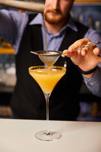 cropped view of bartender pouring cocktail while holding shaker near sieve and margarita glass