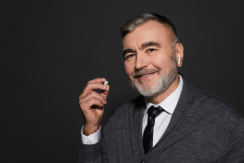 senior man holding earphone while listening music and smiling at camera isolated on dark grey