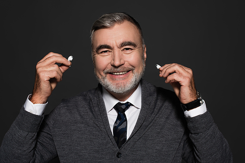 happy senior man in jumper and tie holding earphones isolated on dark grey