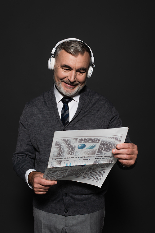 happy senor man in jumper, tie and headphones reading newspaper on dark grey