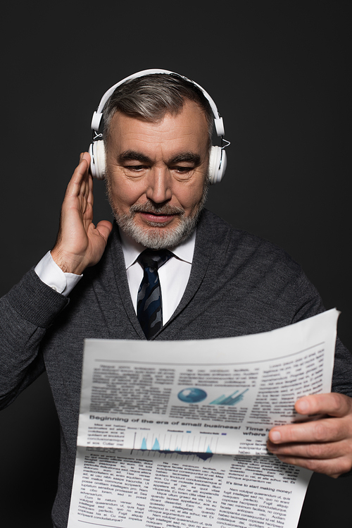 bearded senior man in headphones reading newspaper isolated on dark grey