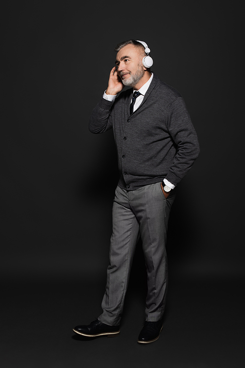 full length view of senior man in stylish clothes listening music in headphones on dark grey