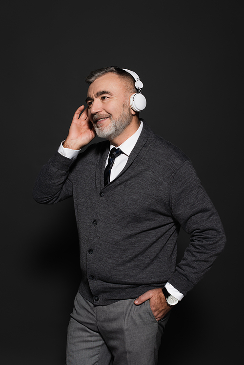 senior stylish man standing with hand in pocket while listening music in headphones on dark grey