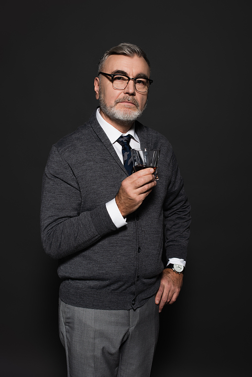 stylish and confident senior man standing with hand on hip and glass of whiskey on dark grey