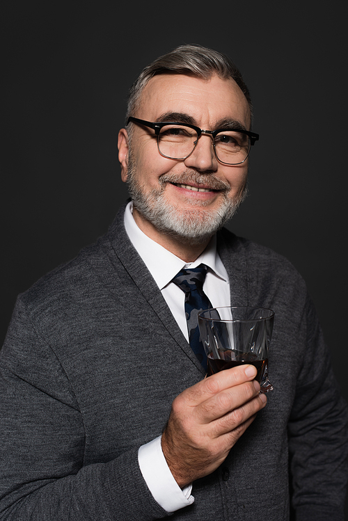 positive senior man in eyeglasses holding glass of whiskey while  isolated on dark grey