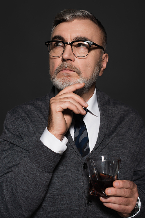 thoughtful senior man with glass of whiskey holding hand near face while looking away isolated on dark grey