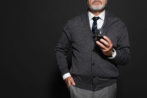cropped view of stylish man with glass of whiskey standing with hand in pocket on dark grey
