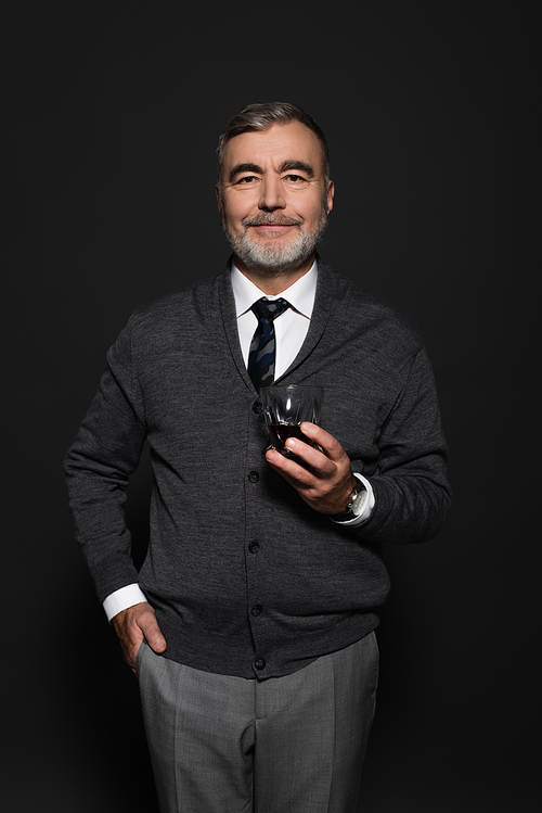 grey-haired man standing with glass of whiskey and hand in pocket while smiling at camera on dark grey