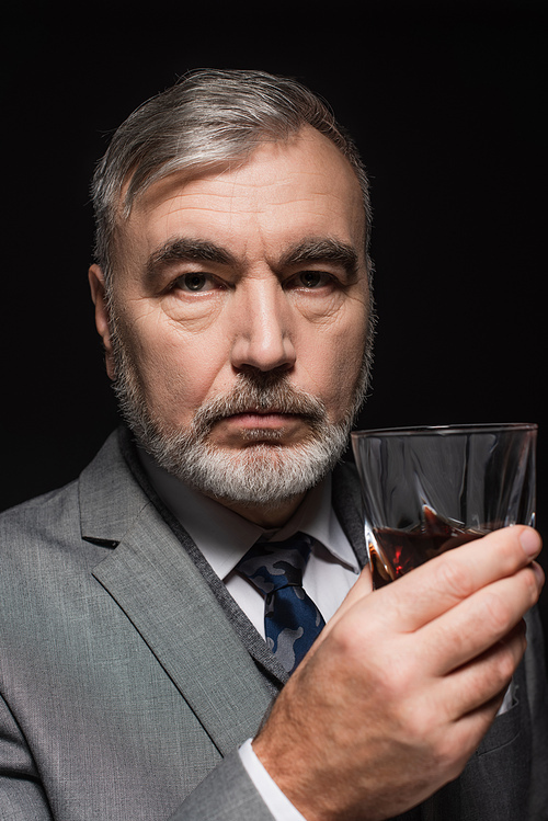 senior bearded man in blazer and tie holding glass of whiskey isolated on black
