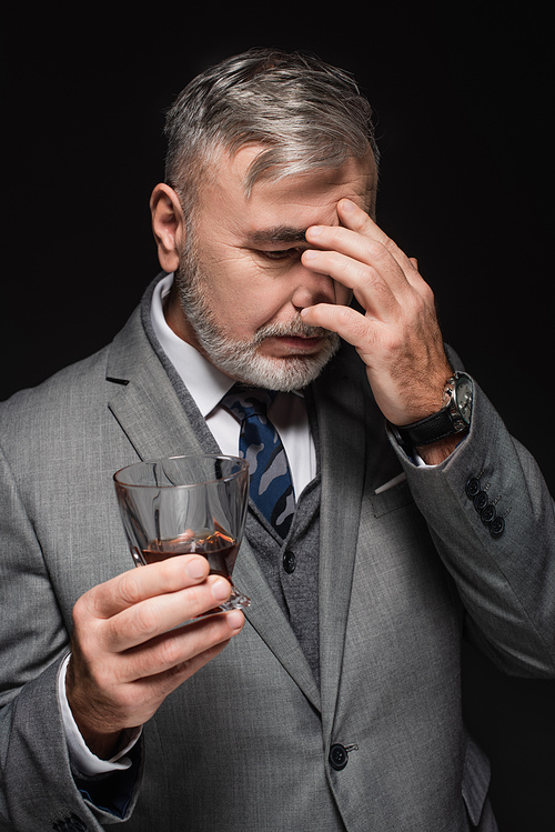 exhausted senior man holding glass of whiskey while suffering from headache isolated on black