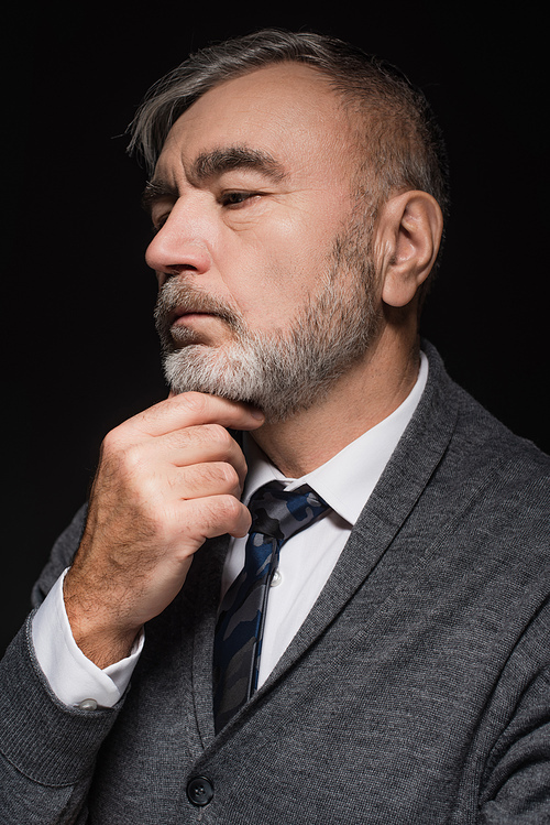thoughtful senior man in grey jumper and tie holding hand near chin isolated on black