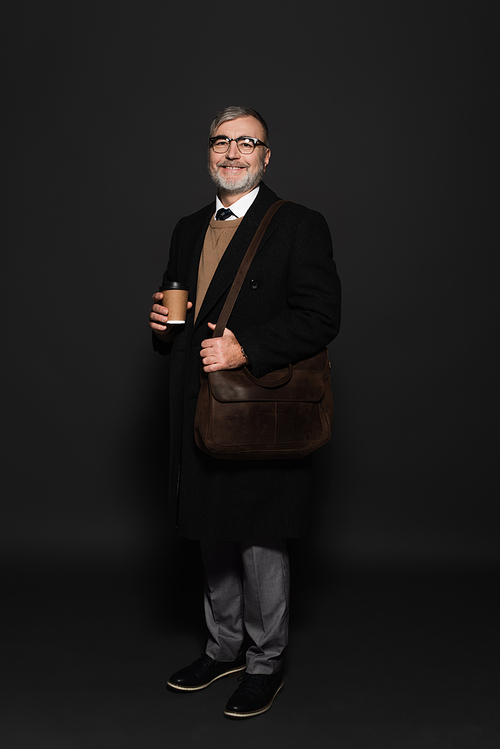 full length view of pleased senior man in black coat standing with leather bag on dark grey