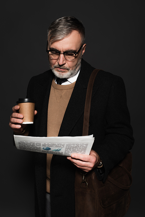 trendy senior man with leather bag and paper cup reading newspaper isolated on dark grey