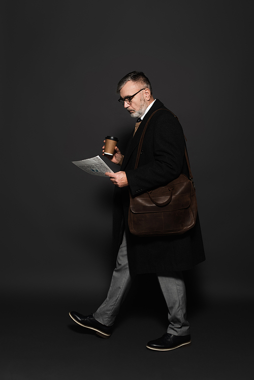full length view of trendy senior man with coffee to go and newspaper walking on dark grey