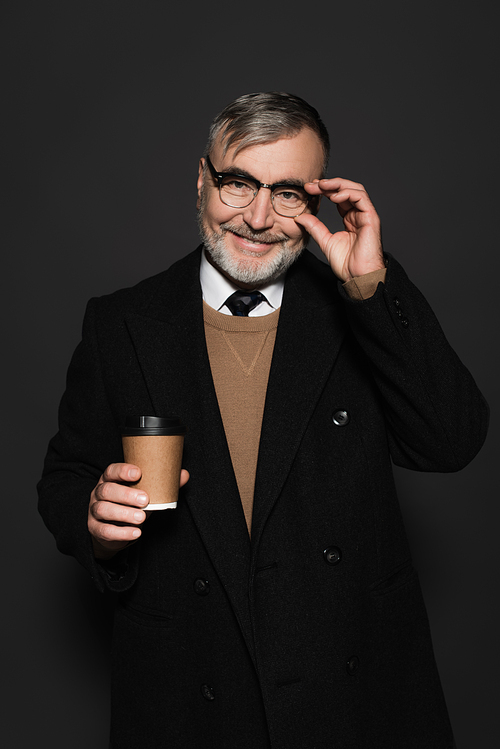 happy man in stylish coat holding paper cup and adjusting eyeglasses isolated on dark grey