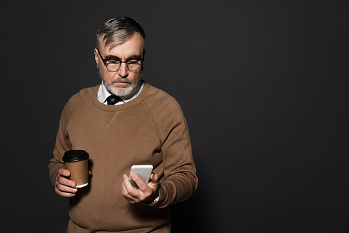 elderly man with coffee to go looking at mobile phone on dark grey