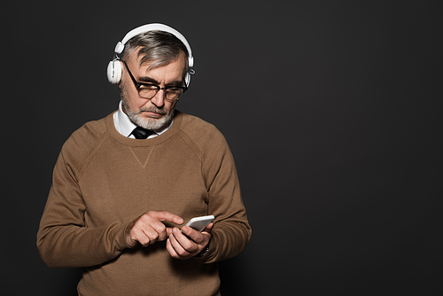 senior man in beige jumper and headphones pointing at mobile phone on dark grey