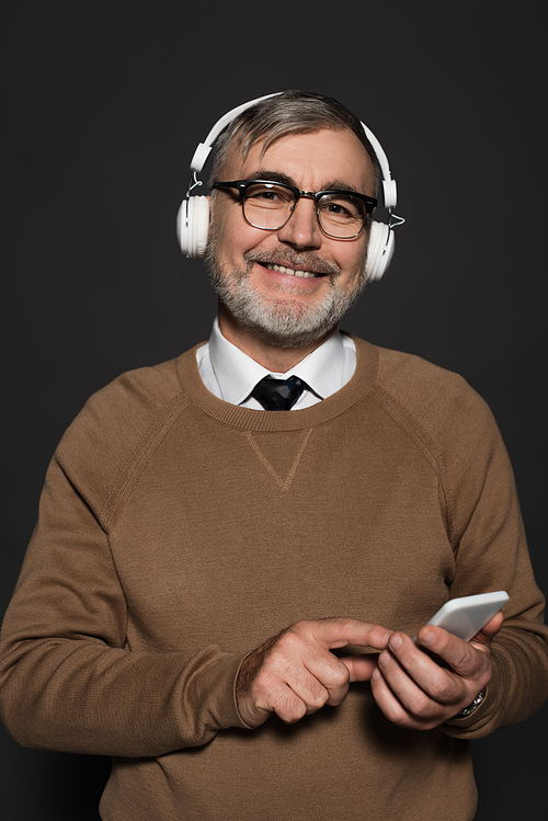 cheerful senior man in headphones  while holding smartphone isolated on black